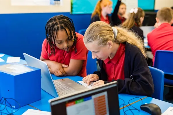 Two primary school girls collaborating on a code-breaking challenge on a laptop with a cipher safe on the desk