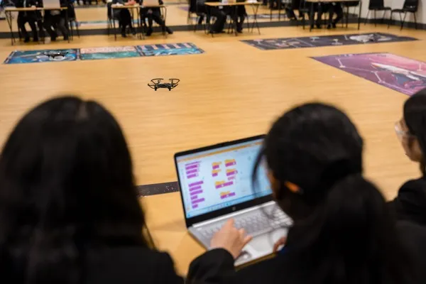 Students coding drone flight paths on a laptop while their drone flies over challenge mats in a school hall