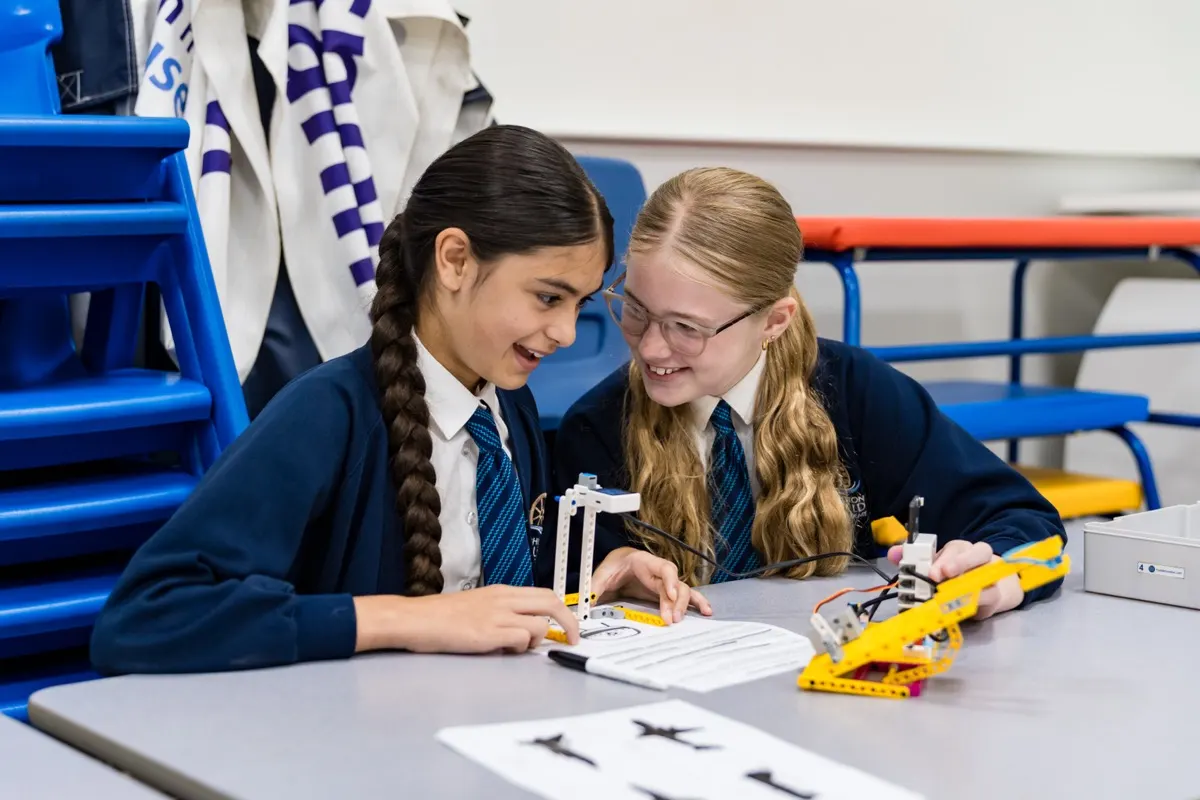 Two girls in school uniform smiling as they collaborate on a LEGO SPIKE robotics project during a STEM workshop