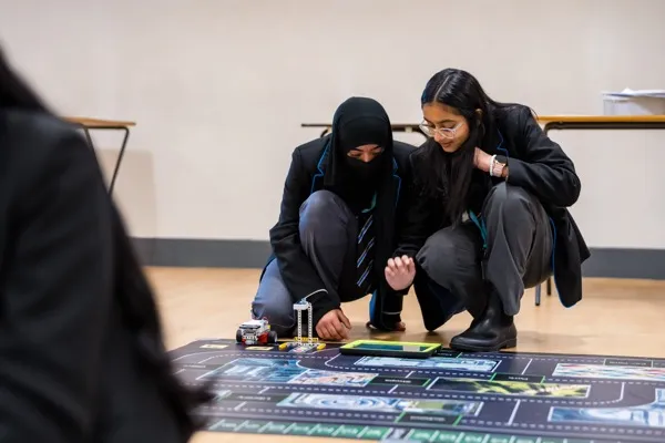 Two students testing their autonomous vehicle robot on the road mat with an iPad nearby