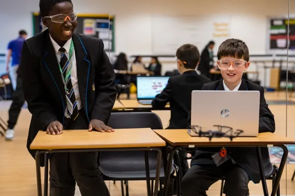 Two secondary students wearing safety goggles, one coding at a laptop with a drone on the desk