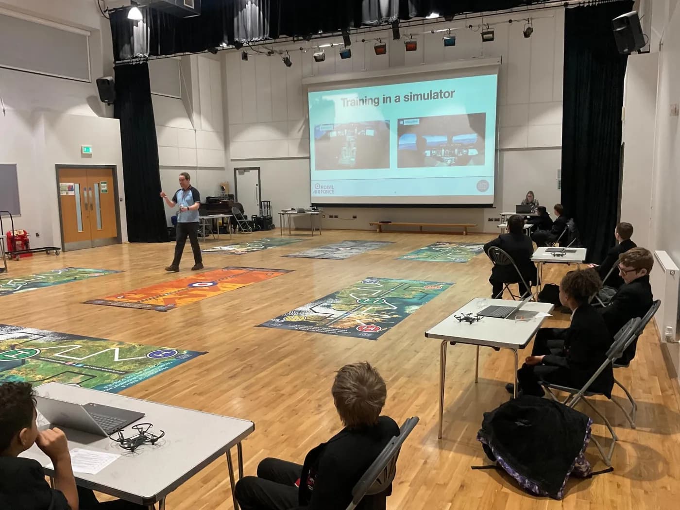 Instructor leading a Drone Coding session from the front of a hall with students in pairs at tables around the perimeter and challenge mats on the floor