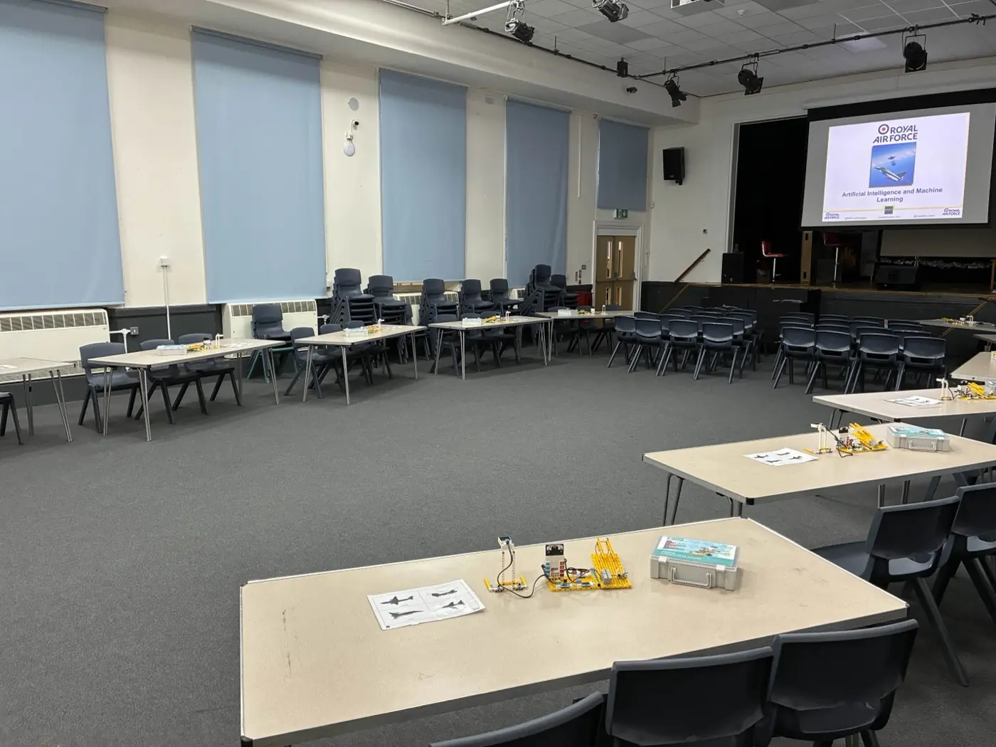 School hall set up for an AI and Machine Learning workshop, with tables carrying robotics kit boxes and rows of chairs facing a screen at the front
