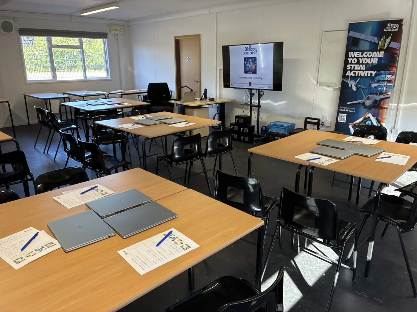 A classroom set up for Cybersecurity & Cryptography, with desks in pairs, laptops on every desk, RAF Youth STEM branding at the front and a projector ready to display the workshop