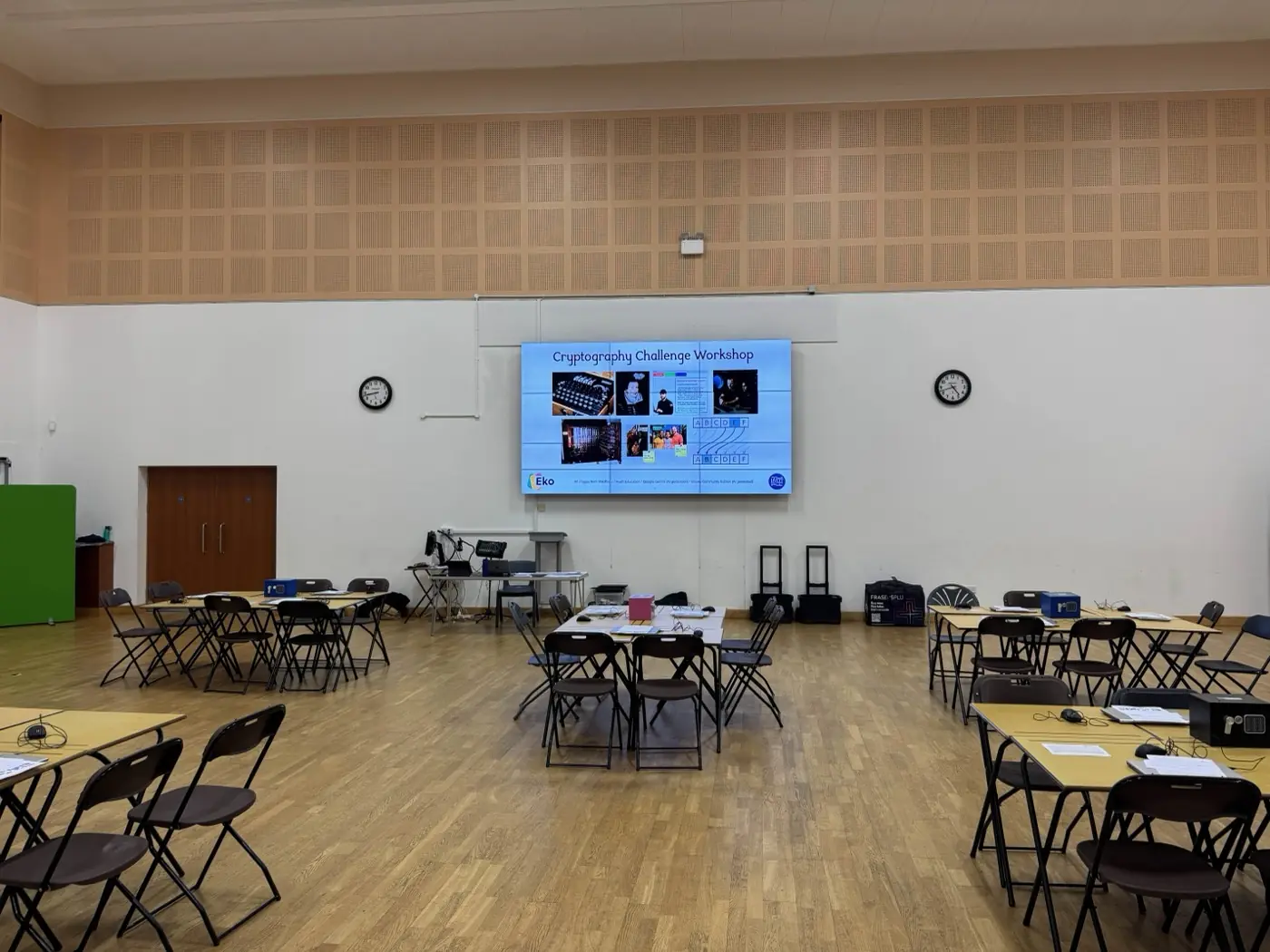 School hall set up for a Cybersecurity and Cryptography workshop, with exam tables grouped for teams and a screen at the front showing the Cryptography Challenge Workshop title