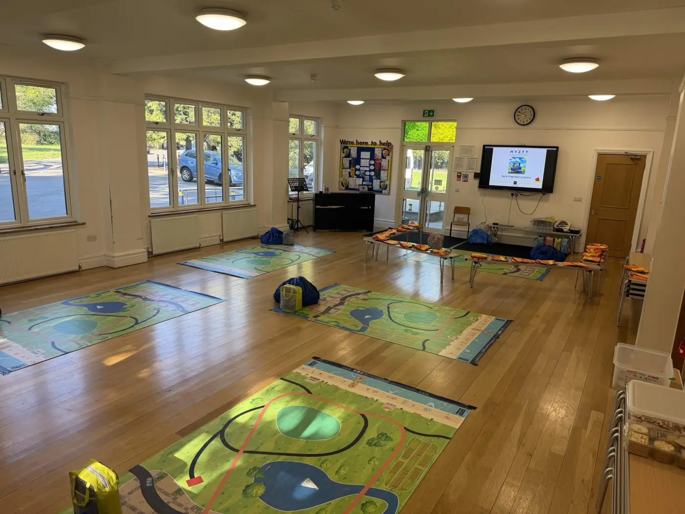 A small school hall set up for Early Engineers Express, with large floor mats laid out on a wooden floor, benches and chairs along the side, and a screen showing the workshop title at the front