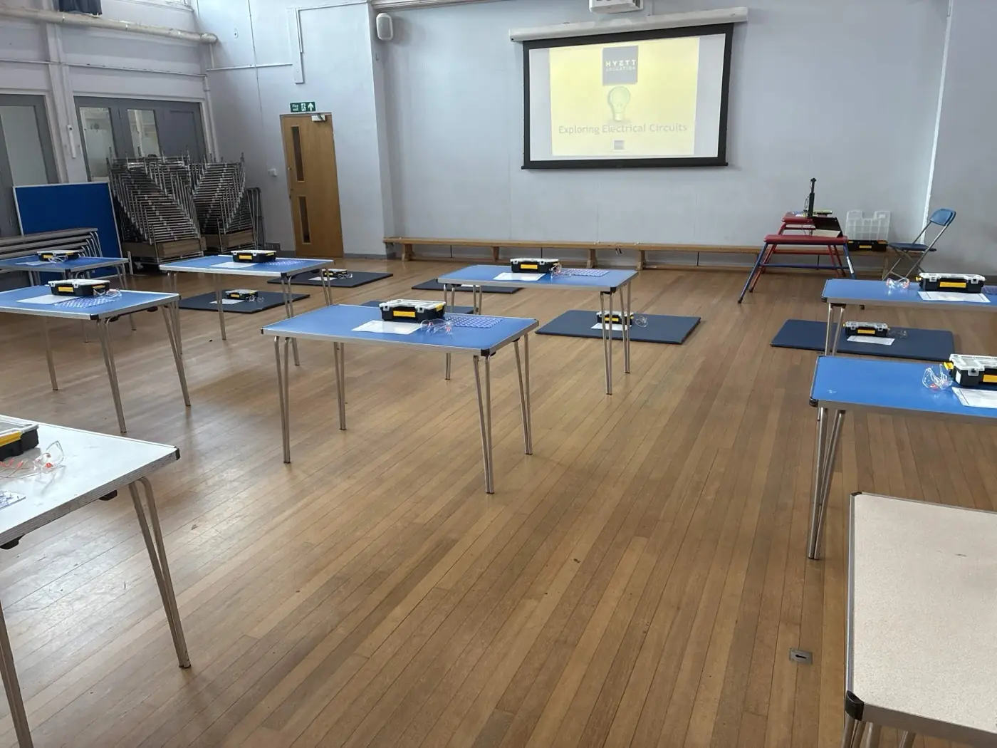 School hall set up for an Electricity and Circuits workshop, with small trestle tables spaced across the floor, each carrying a circuit kit, and a screen at the front